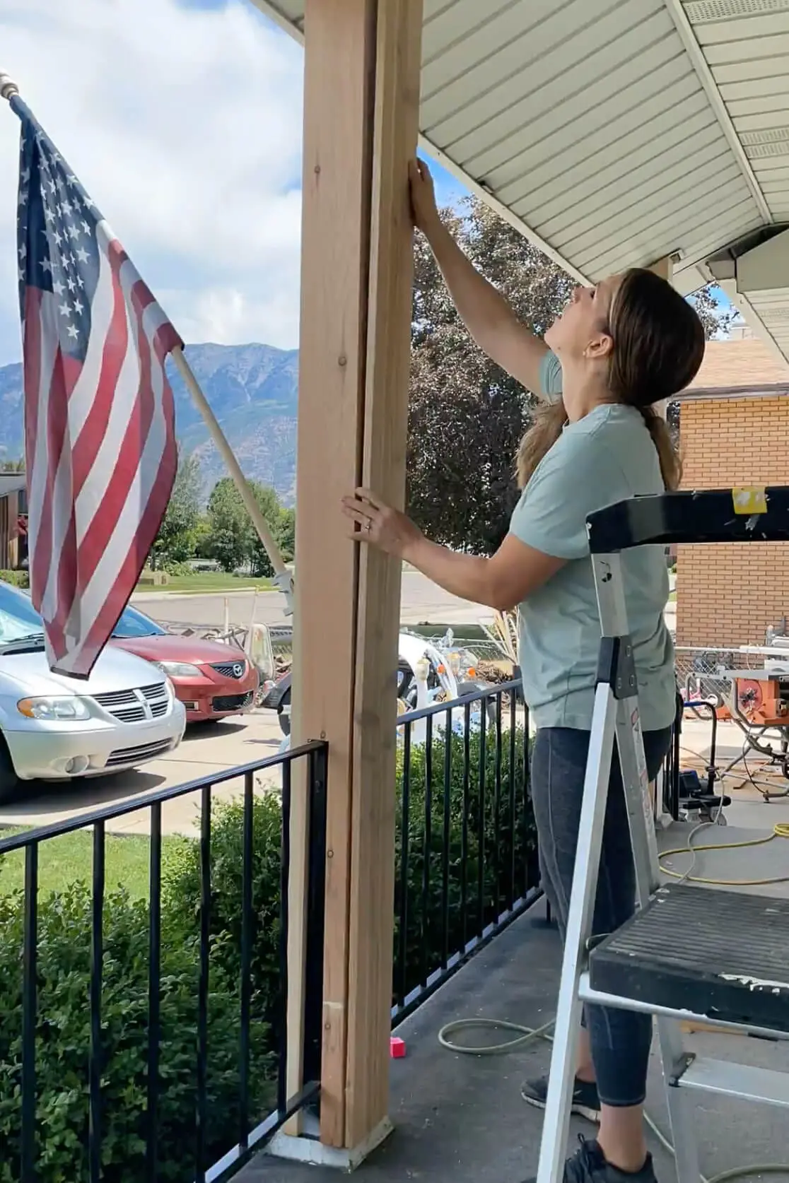 Woman on a step ladder finishing the install of a wooden wrap around a front porch post as part of a DIY makeover to modernize her home's exterior.